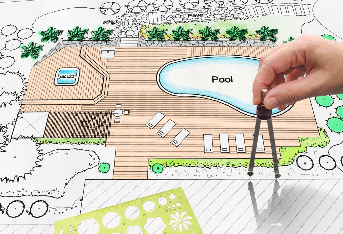 A hand uses a compass over a landscape architectural plan featuring a pool, jacuzzi, lounge chairs, plants, and decking. Templates and design tools are visible on the detailed, colorful drawing.