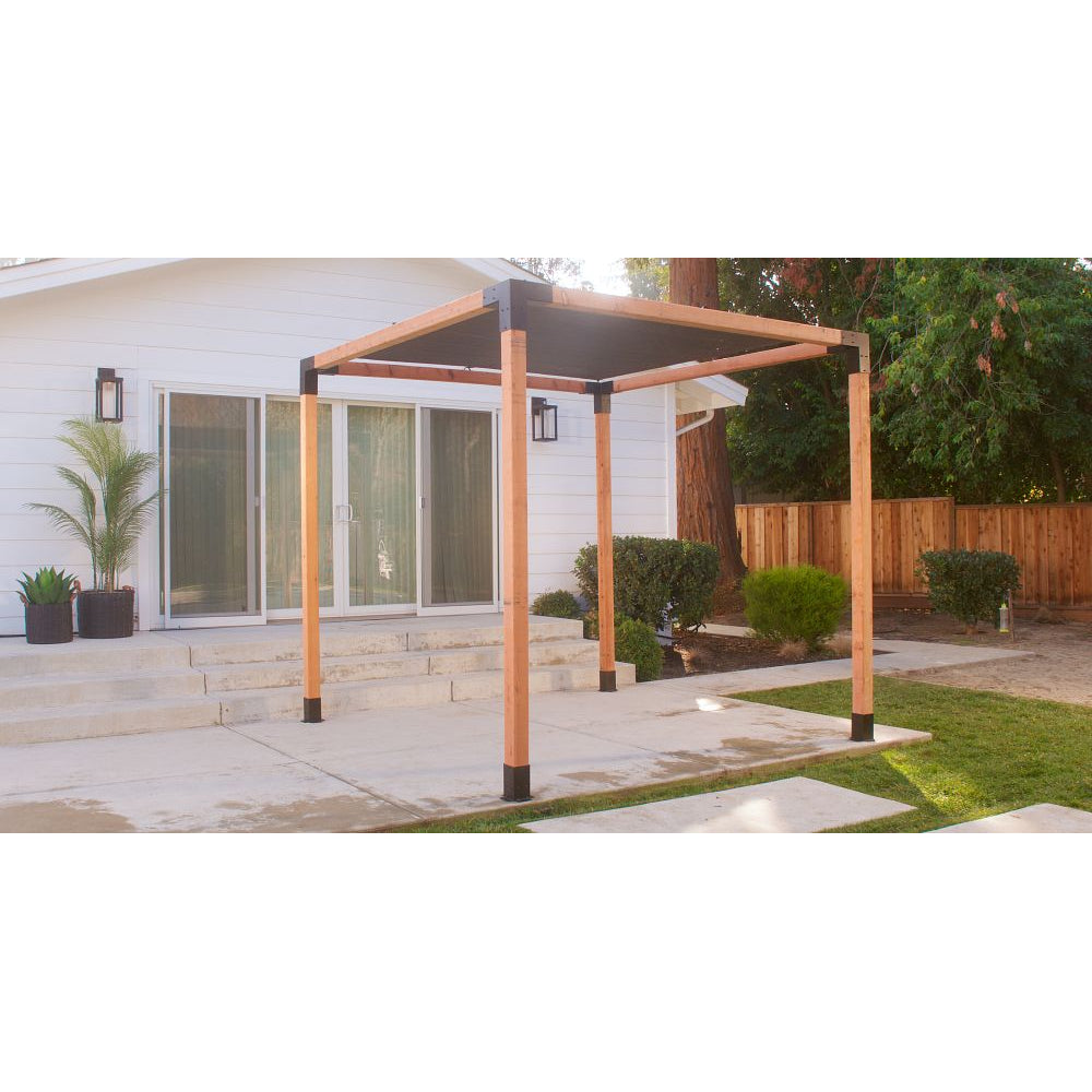 A modern wooden pergola stands on a concrete patio in front of a white house with sliding glass doors, outdoor plants, and a fenced backyard, securely installed using Simpson Strong-Tie® Sage Post Base for 4x4 (black, 2 per pack) for lasting stability.