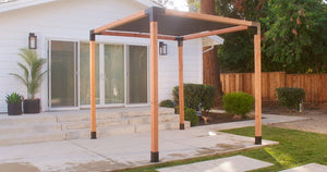 A modern pergola with a black slatted roof, anchored on a concrete patio by Simpson Strong-Tie® Sage Base for 6x6, Black (2 per Pack) hardware, stands in front of a white house with sliding glass doors, surrounded by greenery and a wooden fence.