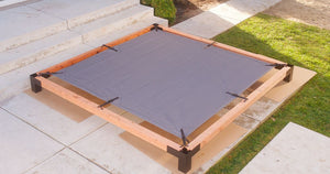 A square wooden frame assembled with Simpson Strong-Tie® Sage Corner Connector for 6x6, Black (2 per Pack), holds a gray fabric sheet above patio tiles near a grassy area. The corrosion-resistant pergola hardware and galvanized steel connectors secure the structure beside steps leading up to a small porch.