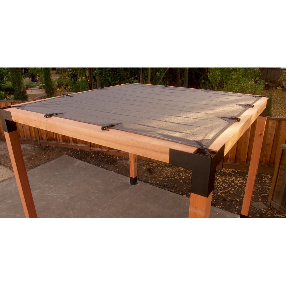 A Sage Sunshade 8x8, Gray by Simpson Strong-Tie® is tightly secured at the corners atop a wooden pergola frame with sturdy ZMAX galvanized steel connectors, standing on a concrete patio in a fenced backyard bordered by green trees.