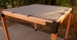 A Sage Sunshade 8x8, Gray by Simpson Strong-Tie® is tightly secured at the corners atop a wooden pergola frame with sturdy ZMAX galvanized steel connectors, standing on a concrete patio in a fenced backyard bordered by green trees.