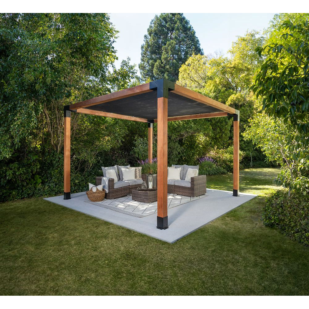 A modern wooden pergola with a Sage Sunshade 8x8 in gray, built using Simpson Strong-Tie® connectors, shades a wicker outdoor seating set with cushions and pillows, placed on a concrete patio surrounded by lush green trees and grass.