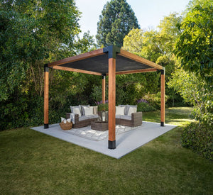 A modern wooden pergola with a Sage Sunshade 8x8 in gray, built using Simpson Strong-Tie® connectors, shades a wicker outdoor seating set with cushions and pillows, placed on a concrete patio surrounded by lush green trees and grass.
