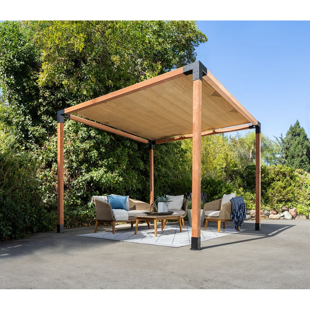 The Simpson Strong-Tie® Sage Sunshade 8x8 Tan pergola kit offers UV protection with its modern wooden design and tan canopy, providing shade for outdoor furniture—such as chairs and a table set on a rug atop the pavement—surrounded by lush green bushes and trees under a clear blue sky.