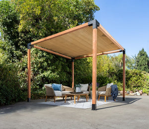The Simpson Strong-Tie® Sage Sunshade 8x8 Tan pergola kit offers UV protection with its modern wooden design and tan canopy, providing shade for outdoor furniture—such as chairs and a table set on a rug atop the pavement—surrounded by lush green bushes and trees under a clear blue sky.