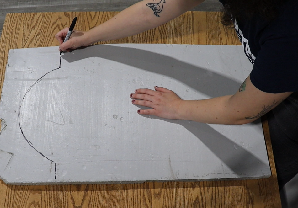 A person with tattoos traces a curved line on a large rectangular piece of foam board placed on a wooden table, using a black marker.