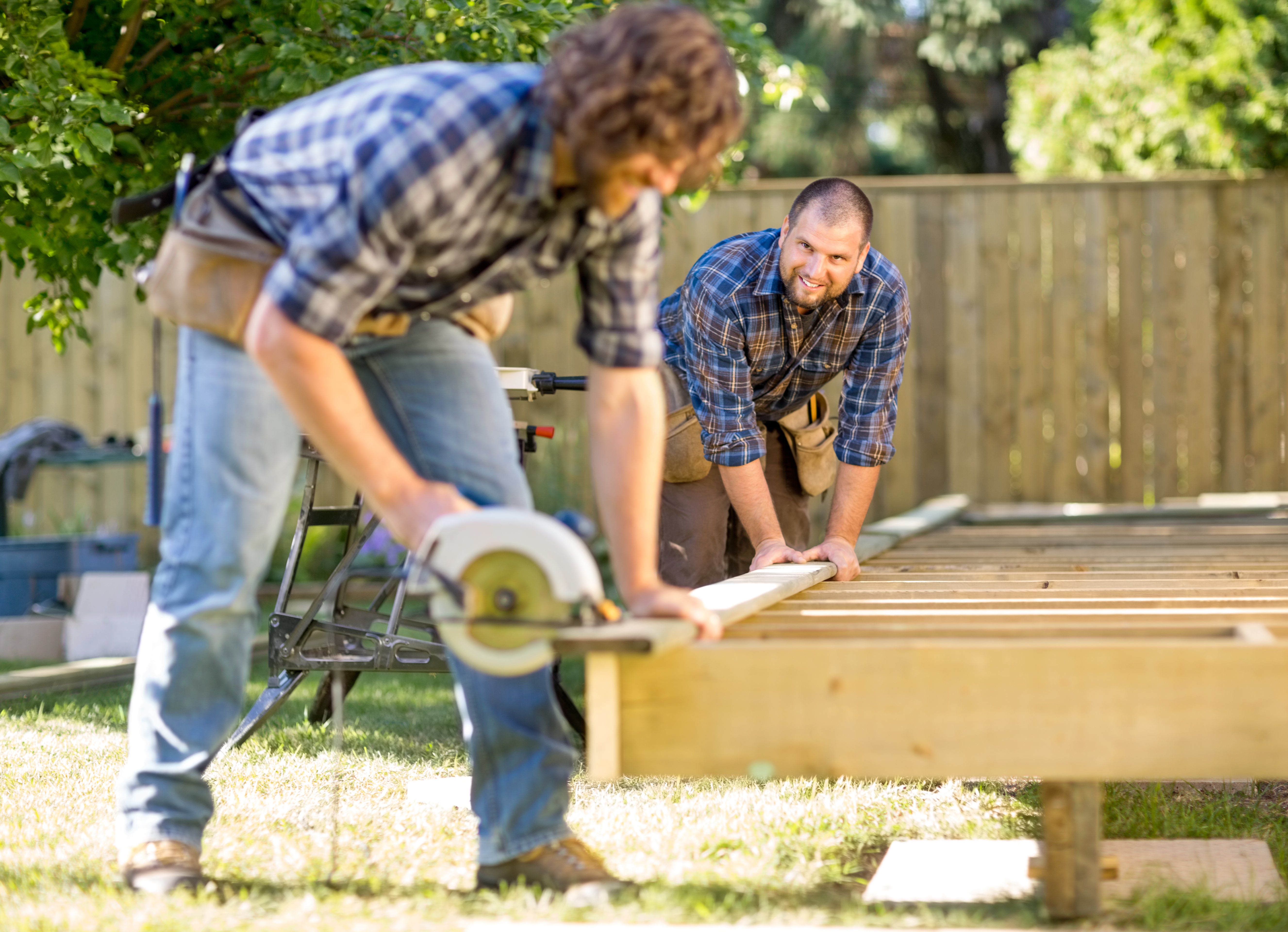 Mid adult carpenter looking at coworker while assisting him in cutting wood with handheld saw
