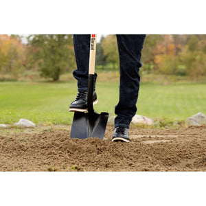 Wearing jeans and black shoes, a person digs into soil on grass with the Garant 8-1/2 Inch Round Point Shovel with Long Wood Handle by Garant Inc, while trees and rocks appear blurred in the background.