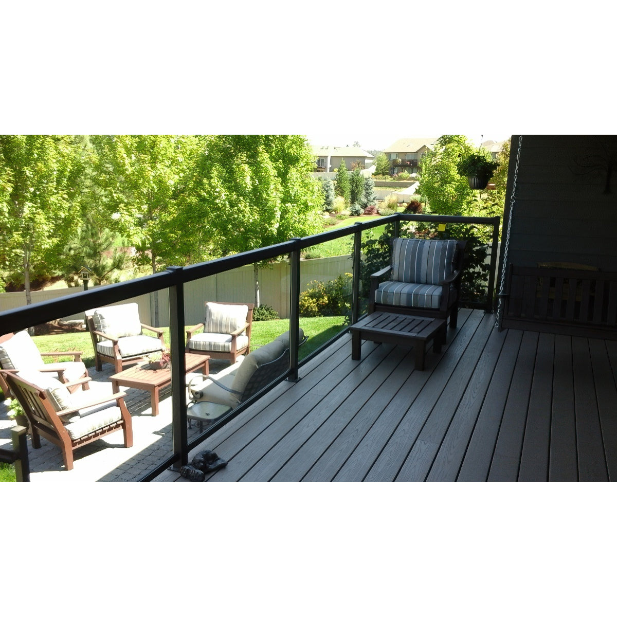 A backyard patio with cushioned chairs and a fire pit is viewed from a wooden deck featuring Regal Rail 36-7/8"X27" Clear Tempered Glass panels (for 42" height), striped armchair, footrest, and lush trees and grass in the background.