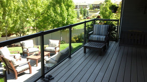 A backyard patio with cushioned chairs and a fire pit is viewed from a wooden deck featuring Regal Rail 36-7/8"X27" Clear Tempered Glass panels (for 42" height), striped armchair, footrest, and lush trees and grass in the background.