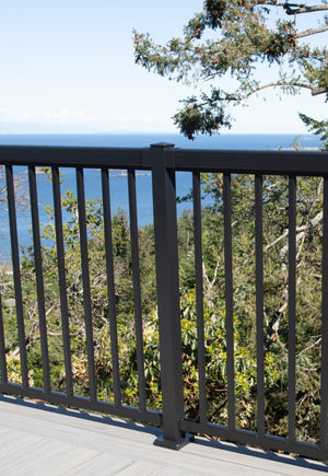 A Regal Rail 6' Straight Pickets, Textured Black (FOR 36") metal railing by Regal Rail lines the deck, overlooking green trees and blue ocean beneath a clear sky.