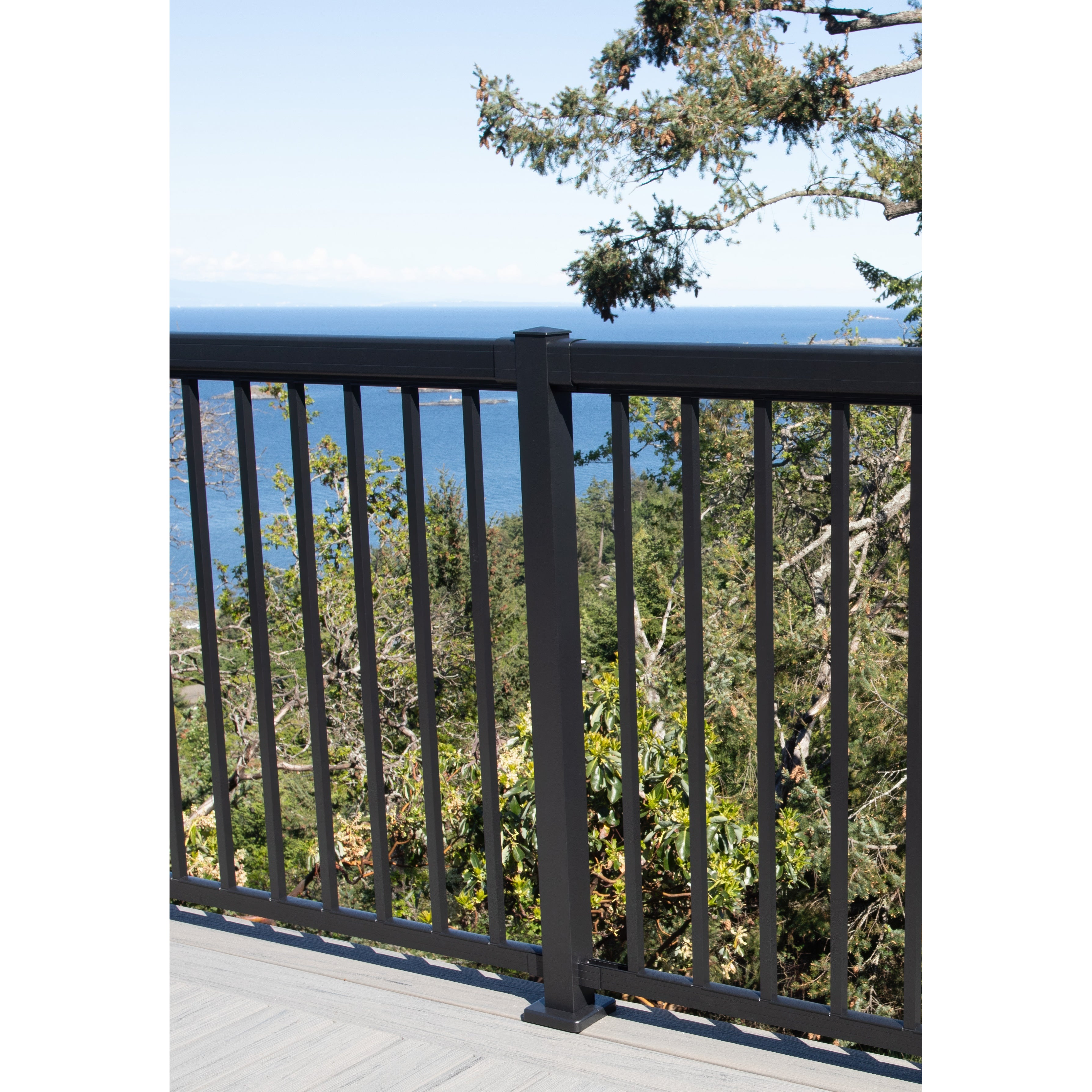 A Regal Rail End Post, Textured Black (FOR 36" HEIGHT), by Regal Rail, stands out on the deck's black Aluminum Railing System, offering corrosion resistance while overlooking green trees and a scenic blue ocean view with a tree branch in the frame.
