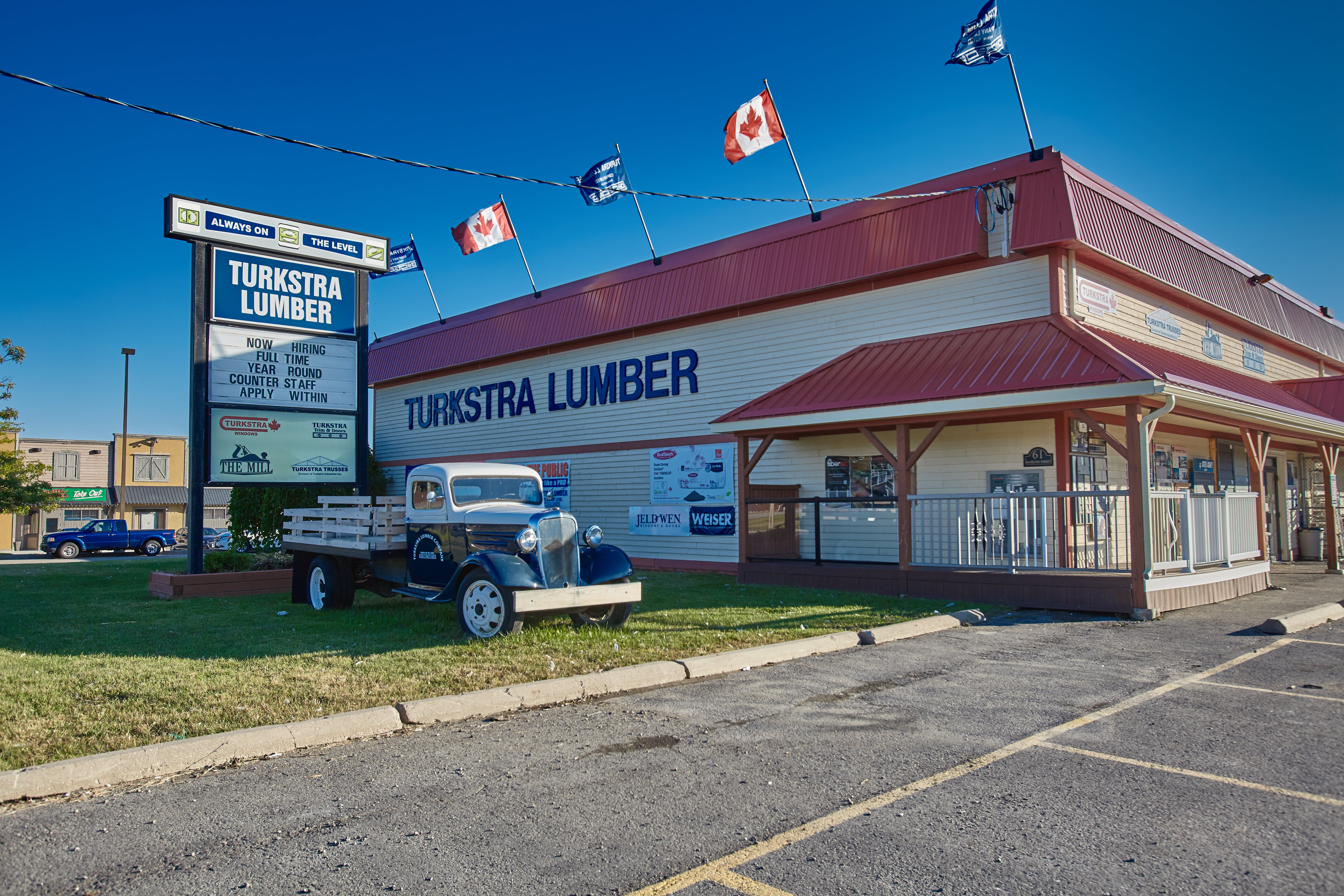 A Turkstra Lumber store with a red roof, a large wall sign, and flags on top. An old-fashioned blue and white truck is parked on the grass near the entrance, and a parking lot is in the foreground.