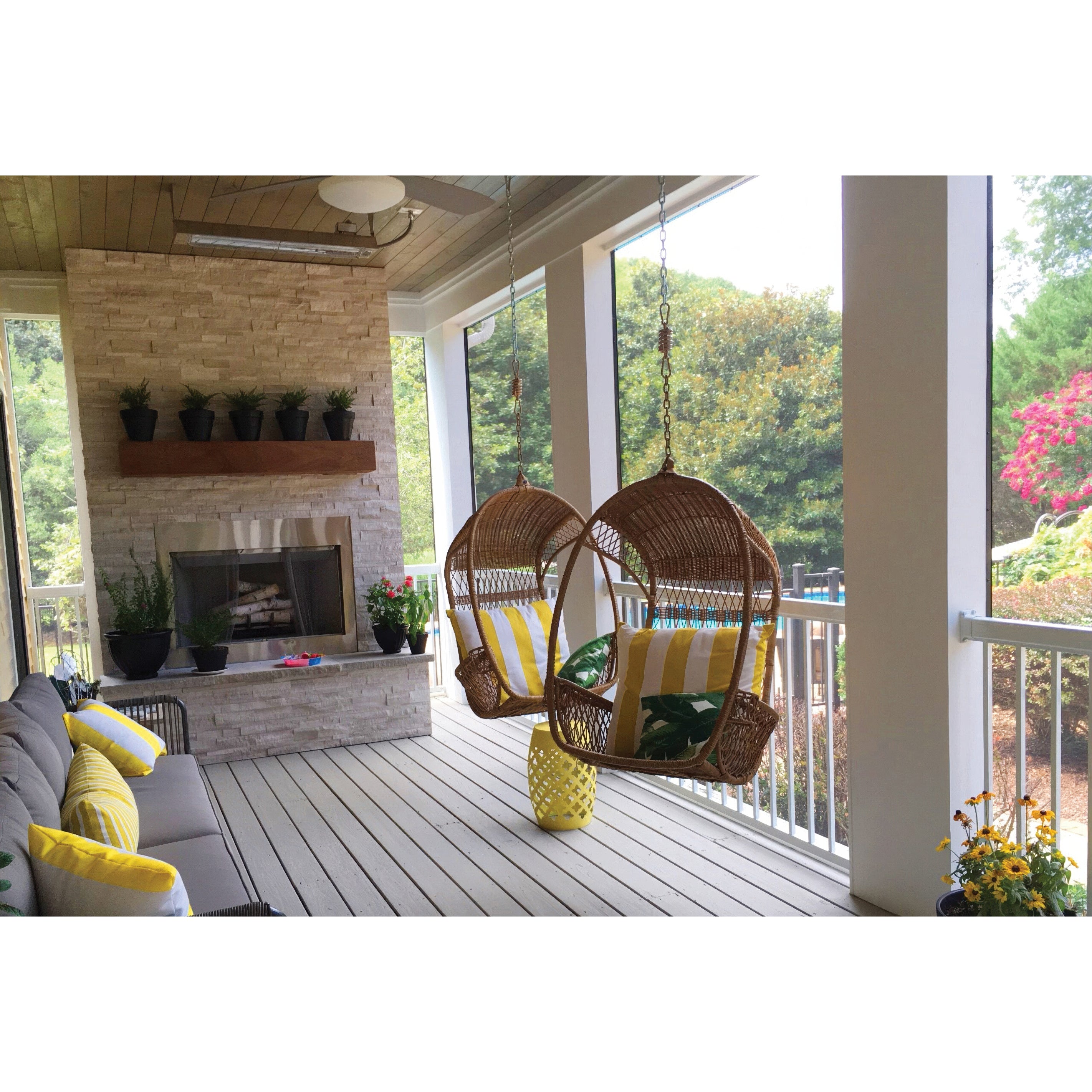 A cozy screened porch features two hanging wicker chairs, a cushioned sofa with yellow-striped pillows, potted plants, a stone fireplace, and Regal Rail 6' Straight Pickets in white framing the view of trees and a garden outside.