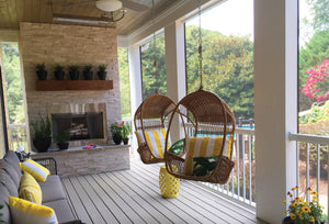 A cozy screened porch with two hanging wicker chairs, a gray sofa, a stone fireplace topped with potted plants, yellow accents, and Regal Rail 8' Straight Pickets in white by Regal Rail edging the space, overlooking a lush green backyard.