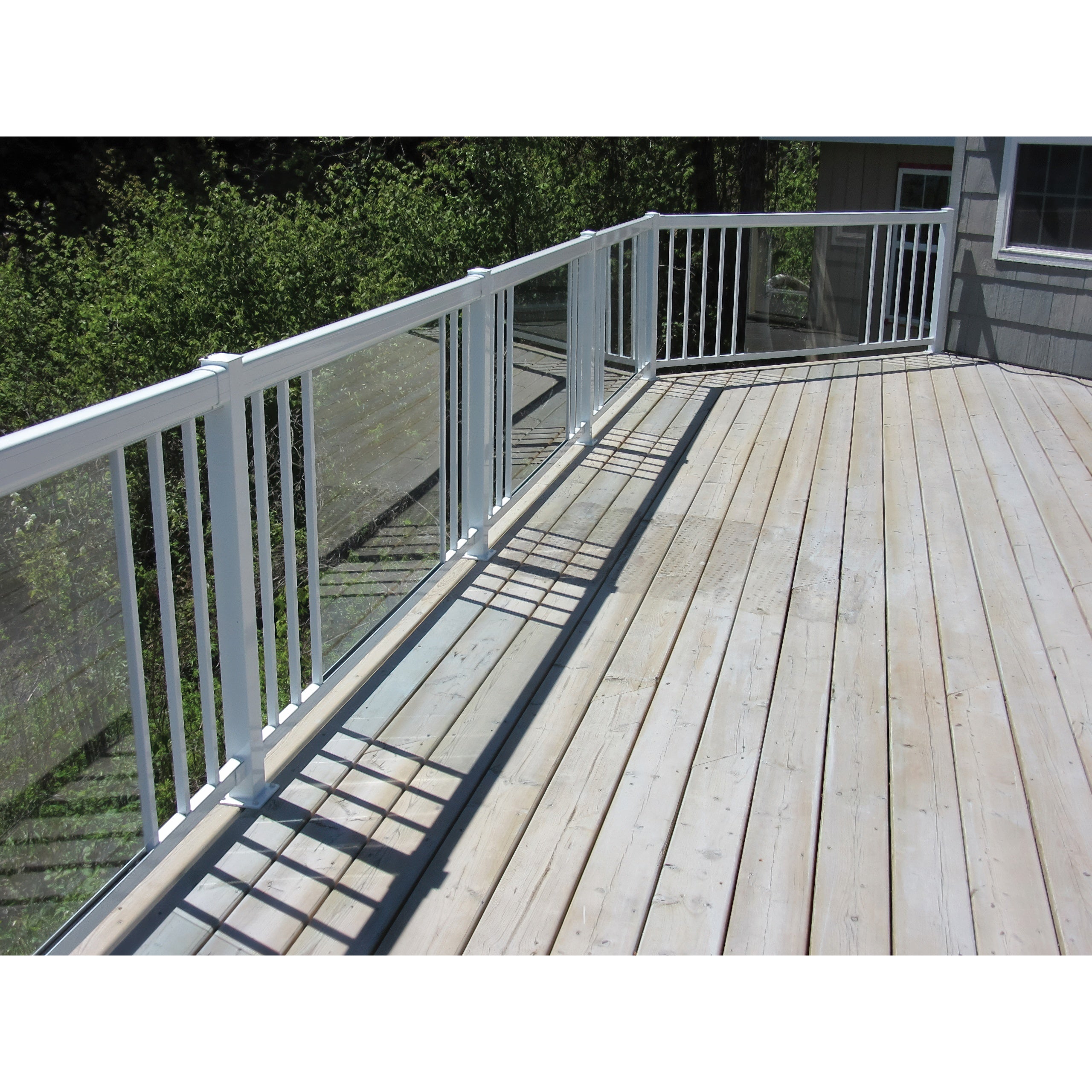 A wooden deck with light planks features a Regal Rail 6' Straight Pickets, White (FOR 42") aluminum railing by Regal Rail, with narrow white pickets and glass panels overlooking greenery. Sunlight casts shadows on the deck.