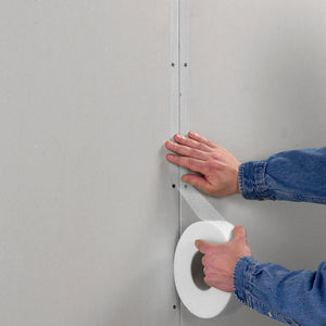 Wearing a denim shirt, a person applies Saint-gobain Adfors 48mm X 500' Fibatape Joint Tape, White to a drywall seam, smoothing the tape with one hand while unrolling it with the other.