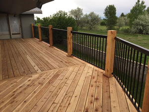 A wooden deck features Regal Rail 6' Straight Pickets, Bronze (FOR 42") by Regal Rail as a railing system, extending from a house and overlooking a grassy yard with trees beneath a cloudy sky.