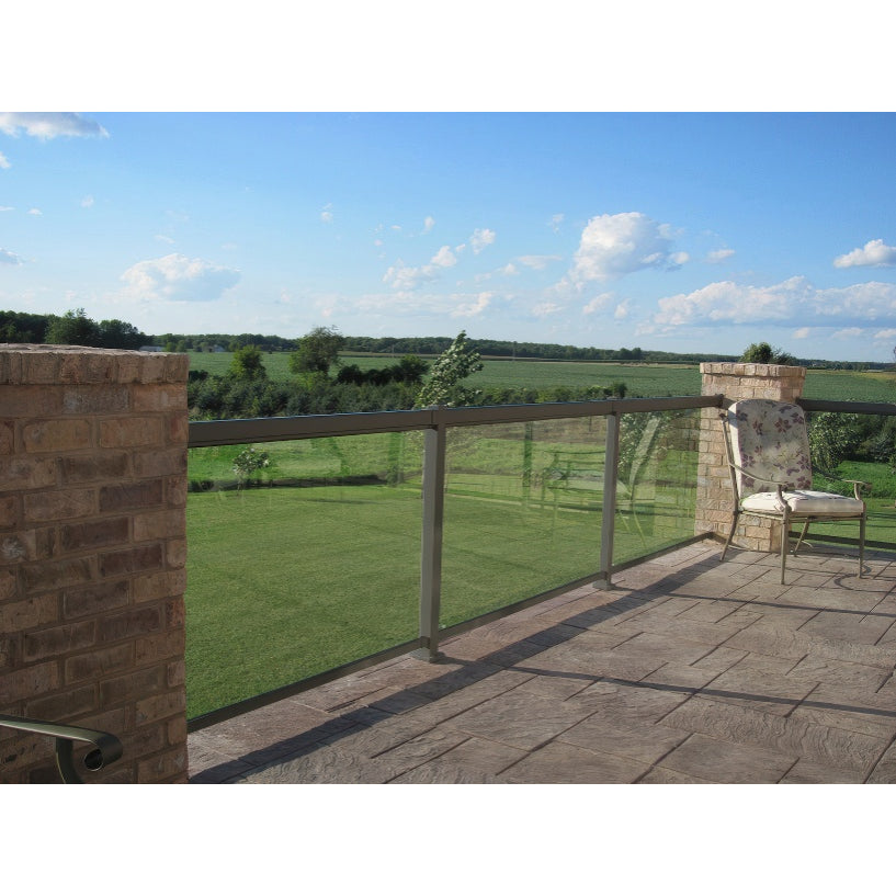 A patio with Regal Rail 6' Straight Pickets, Bronze by Regal Rail, overlooks a green lawn and fields. In the corner sits a chair with floral cushions, while a bright blue sky dotted with clouds completes the scene.