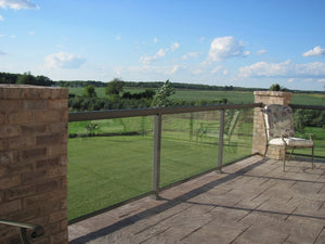 A sunny patio with a stone floor and glass, plus Regal Rail 6' Straight Pickets in Titanium Slate (FOR 42") from Regal Rail, features stylish aluminum railings that overlook green fields under a blue sky for added safety and modern appeal.