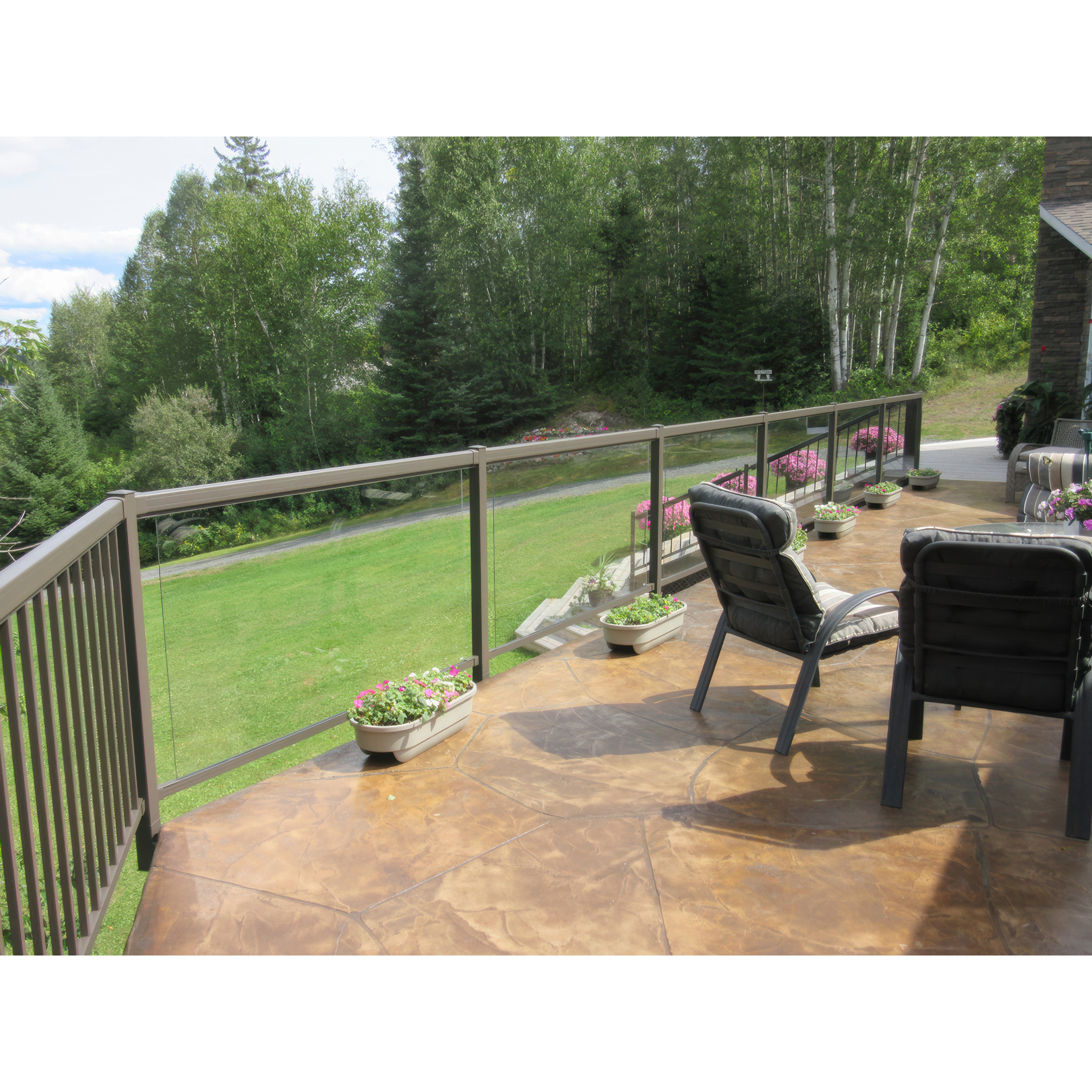 Two cushioned chairs sit on a patio with stamped concrete and flower pots. A Regal Rail 10' Straight Pickets, Bronze (FOR 42") aluminum railing overlooks a green lawn, trees, and a distant forest.