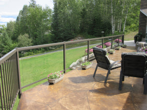 Two cushioned chairs sit on a patio with stamped concrete and flower pots. A Regal Rail 10' Straight Pickets, Bronze (FOR 42") aluminum railing overlooks a green lawn, trees, and a distant forest.