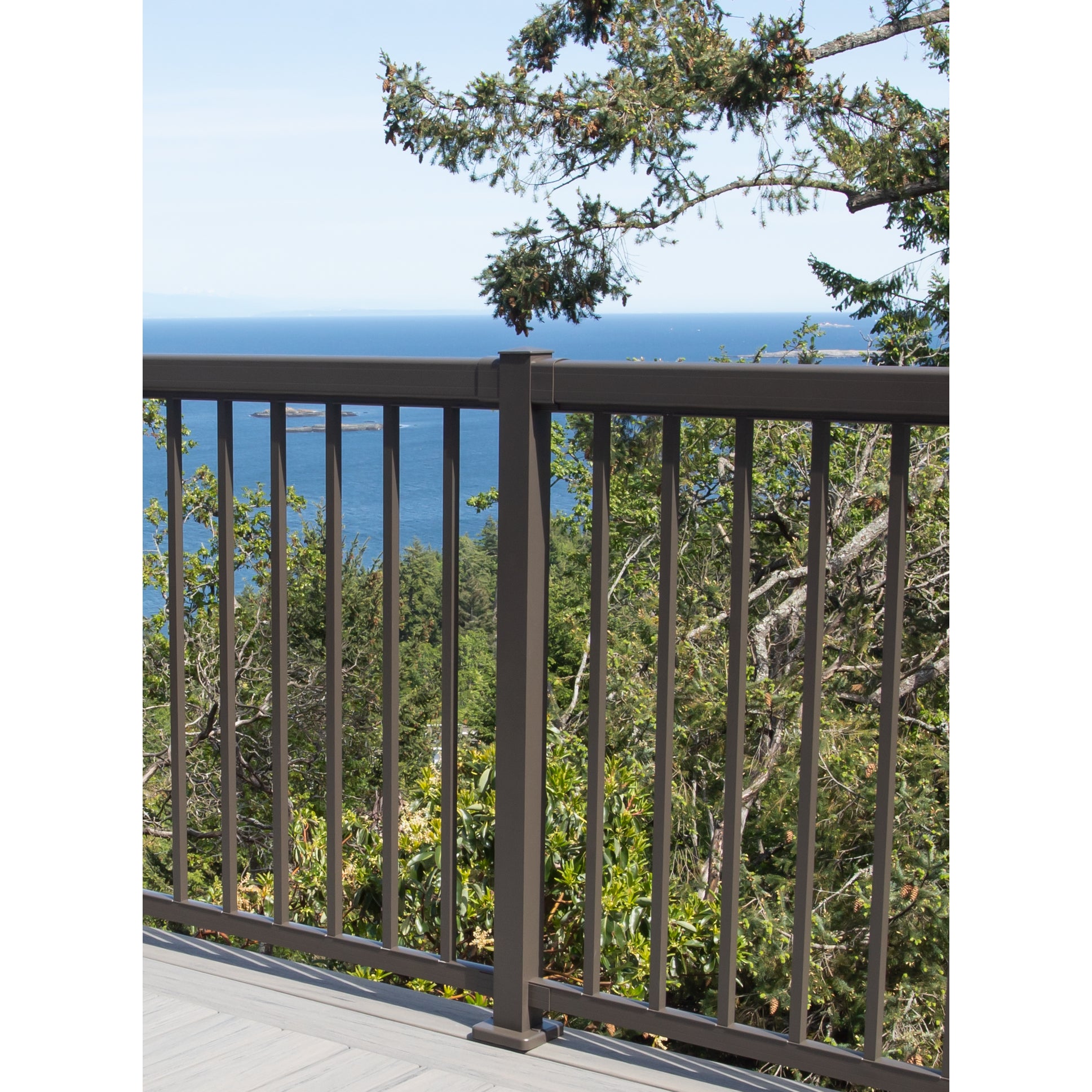 The Regal Rail 8' Straight Pickets in bronze by Regal Rail border the deck, overlooking green trees and a blue ocean. A tree branch extends into view as the UV powder-coated aluminum ensures durability. For use with 42" rails.