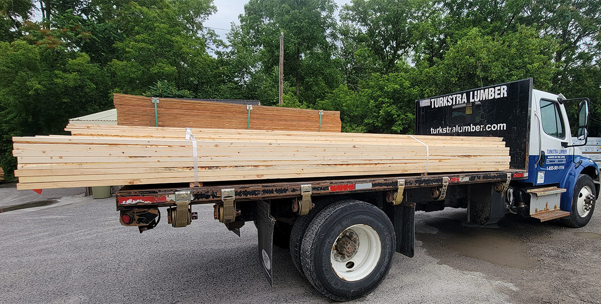 A flatbed truck loaded with stacks of wooden planks and lumber sits parked on a gravel surface, surrounded by trees. The truck has Turkstra Lumber branding on the side.