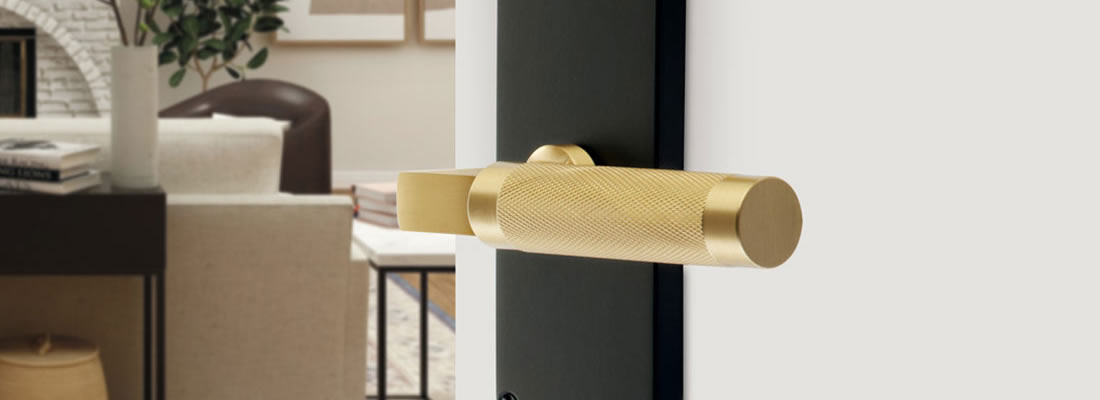 A close-up of a modern gold door handle with a textured grip on a black door, with a living room featuring a sofa and bookshelf visible in the blurred background.