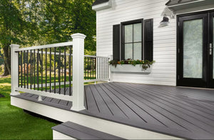 A small gray deck made with Trex Enhance Basics Grooved, Pebble Beach decking is attached to a white house with black shutters and overlooks a grassy yard bordered by trees. The deck’s surface is fade and stain resistant.