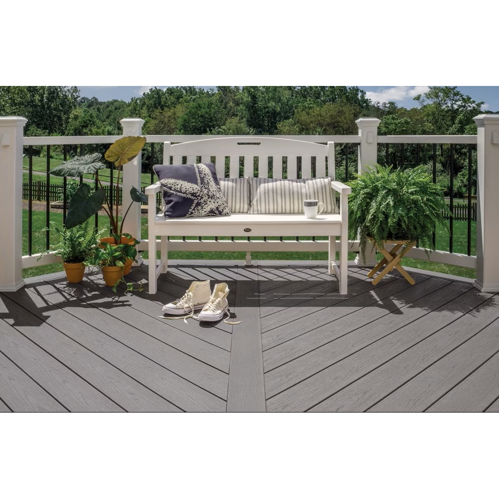 A white bench with cushions and a mug sits on low-maintenance Trex 1" x 12" x 12' Enhance Basics Pebble Beach deck boards. Nearby are potted plants and sneakers, with green trees and a lawn seen past the railing.