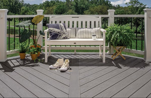 A white garden bench with pillows and a cup sits on Trex Enhance Basics Square 1" x 6" x 20' Pebble Beach decking, surrounded by potted plants, sneakers, a fern on a stand, with lawn and trees visible in the background.