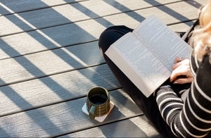 A person in a black and white striped sweater reads a book on sunlit 1" x 6" x 16' Trex Enhance Basics Grooved, Pebble Beach composite decking by Trex. A green coffee mug sits nearby as shadows fall across the deck boards.