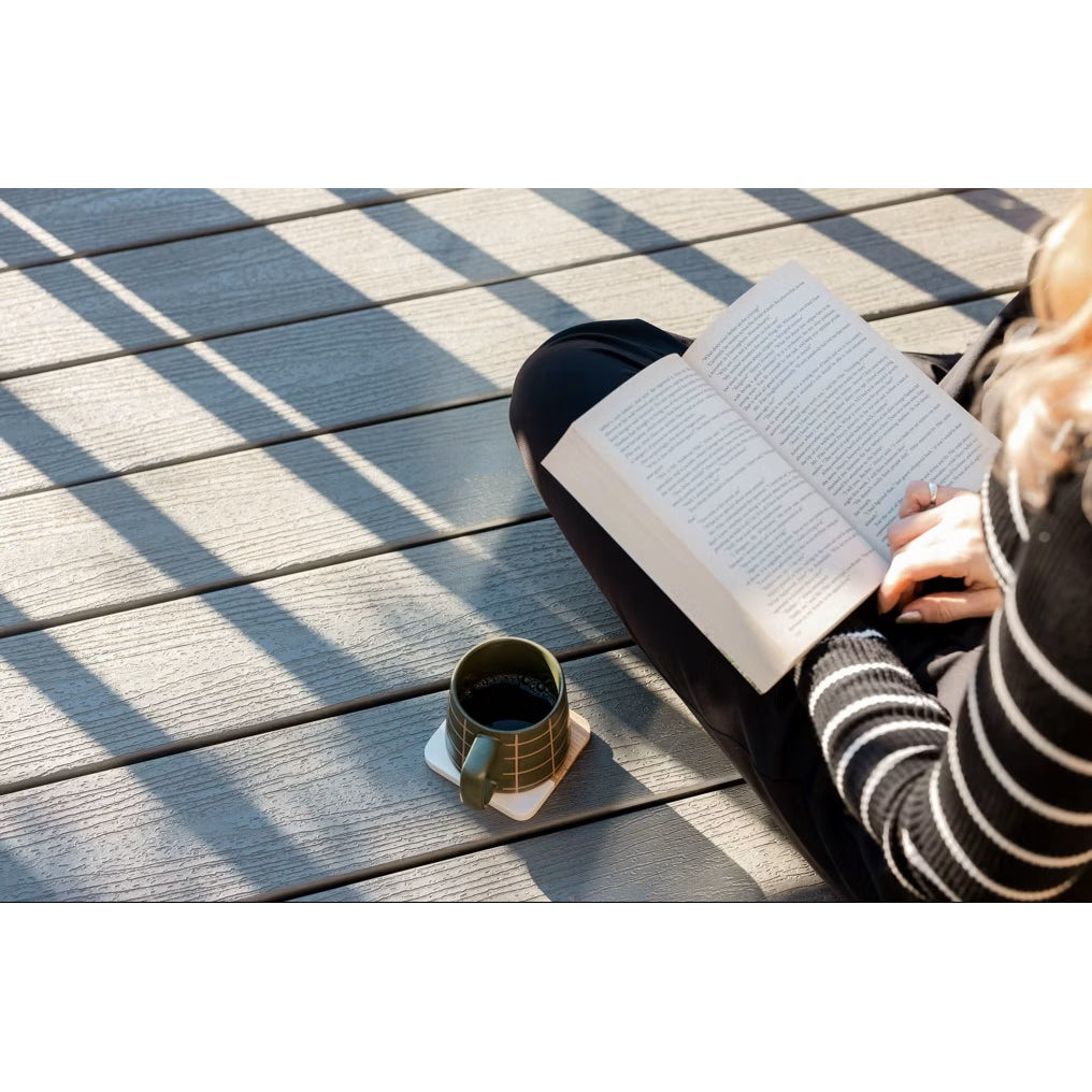 On a sunny day, someone in a striped sweater relaxes on a deck made of Trex Enhance Basics 1" x 12" x 12' Pebble Beach decking, enjoying a book and coffee. Sunlight casts intricate shadows across the durable Trex boards.