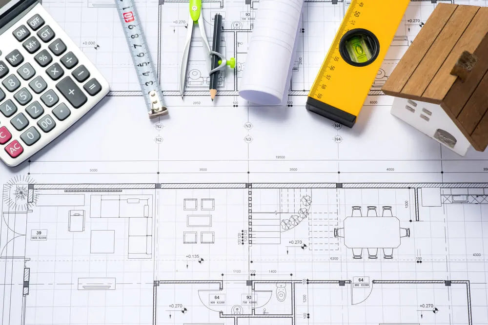 A floor plan blueprint with a calculator, tape measure, compass, level, pencil, rolled paper, and a small model house on top, representing architectural planning and design.