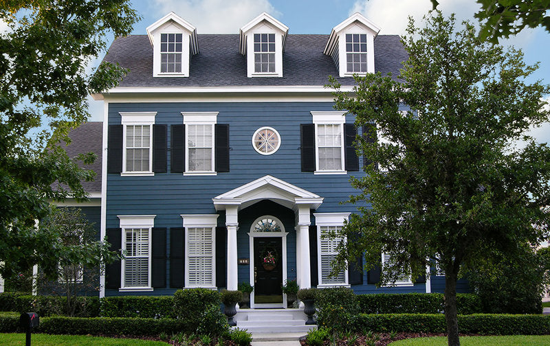 Blue house with white trim and black shutters, surrounded by trees and greenery.