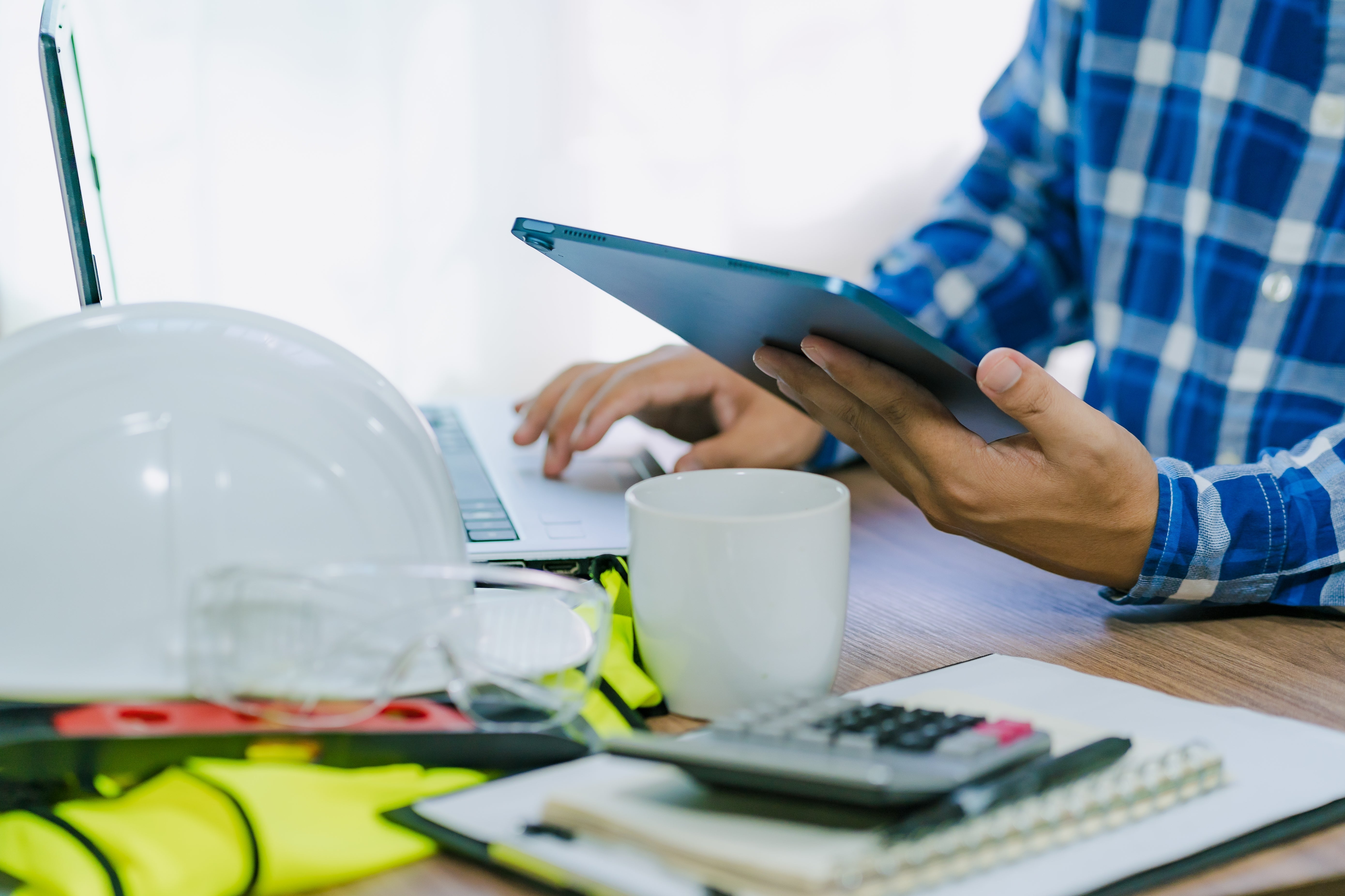 A person in a blue plaid shirt works at a desk with a tablet and laptop. A white hard hat, safety glasses, coffee mug, calculator, and documents are on the desk, suggesting a construction or engineering setting.