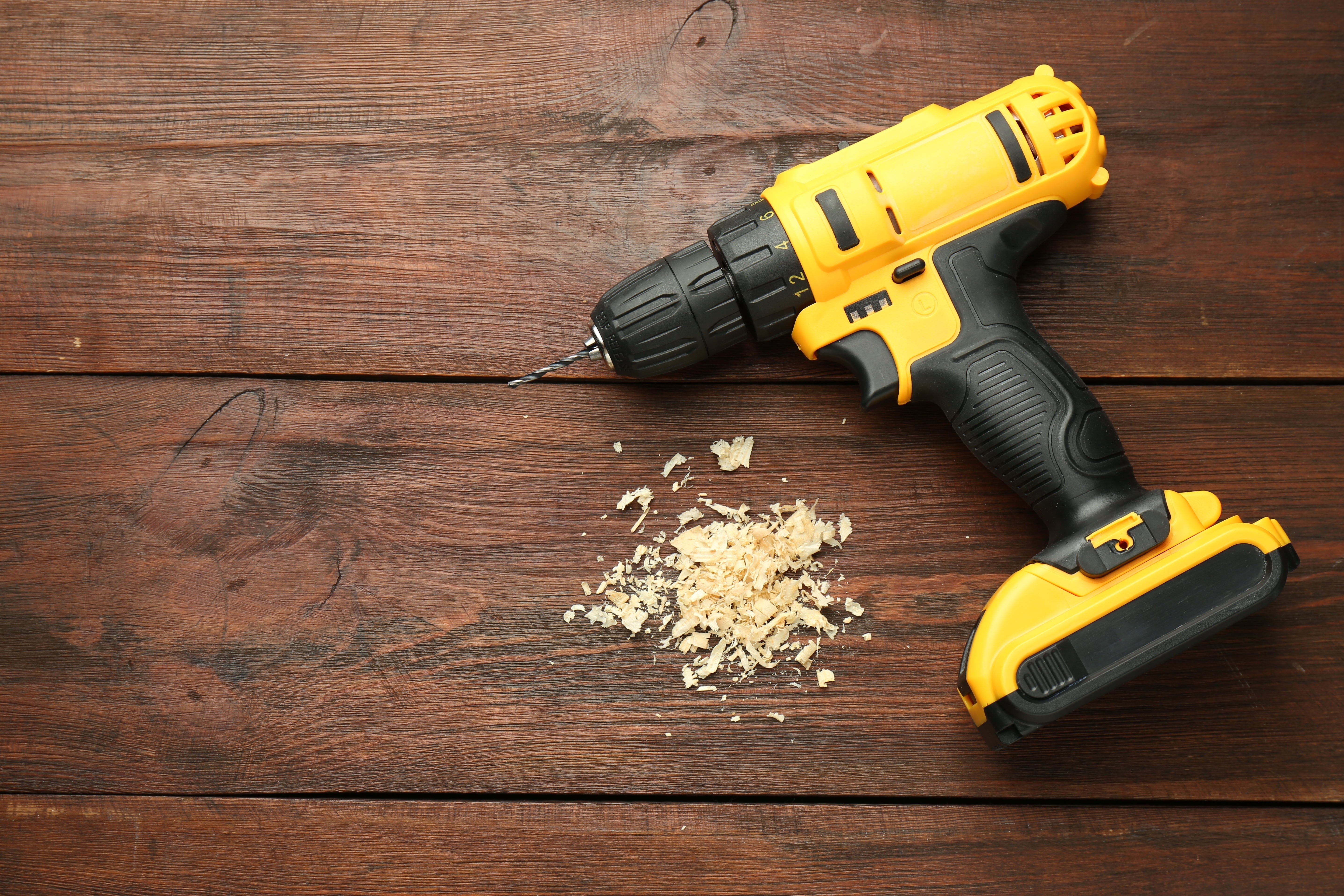A yellow and black cordless power drill with a bit attached lies on a wooden surface next to a small pile of wood shavings.