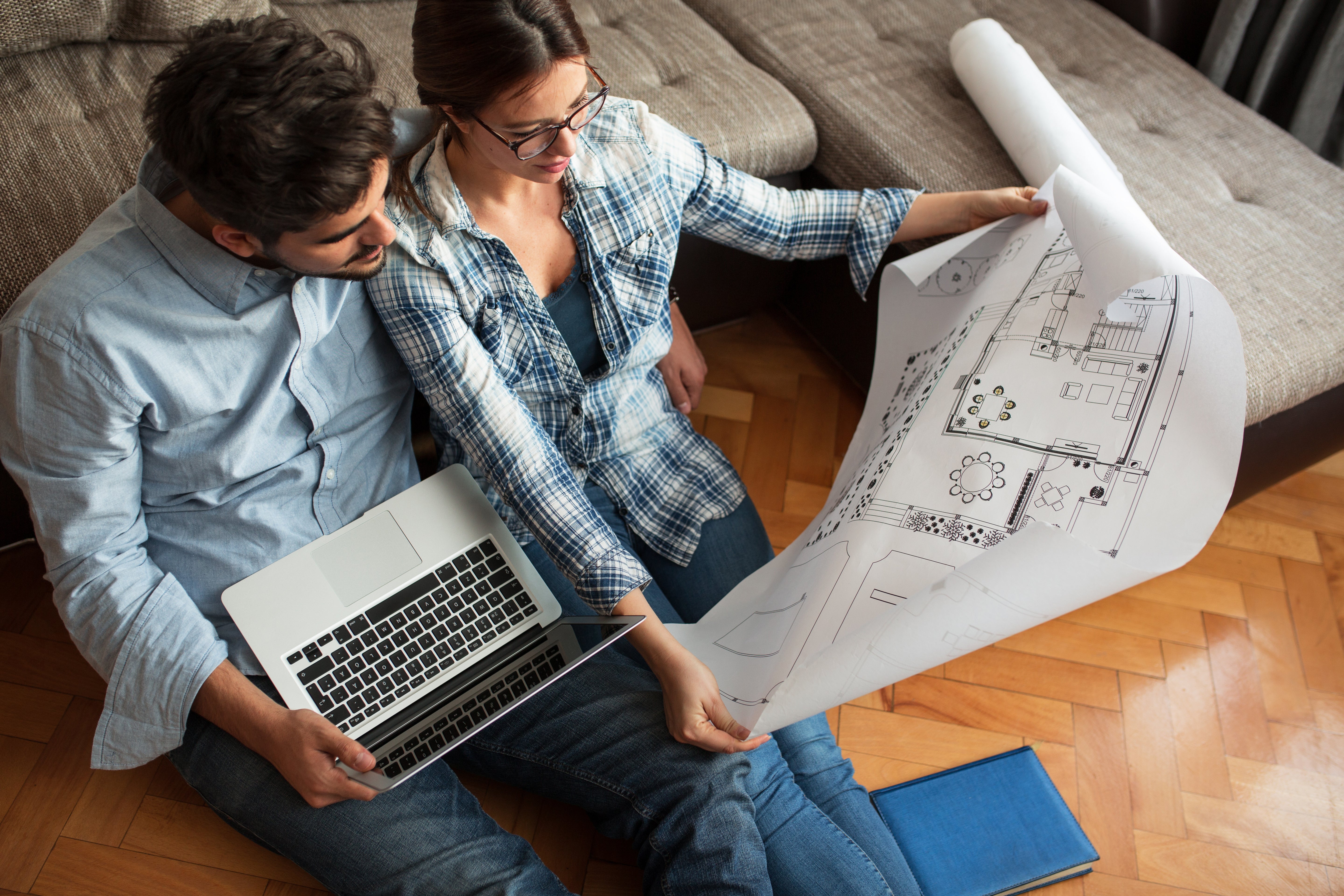 A couple sits on the floor looking at a large architectural blueprint together; one person holds the blueprint while the other uses a laptop. They are sitting near a sofa on a wooden floor.