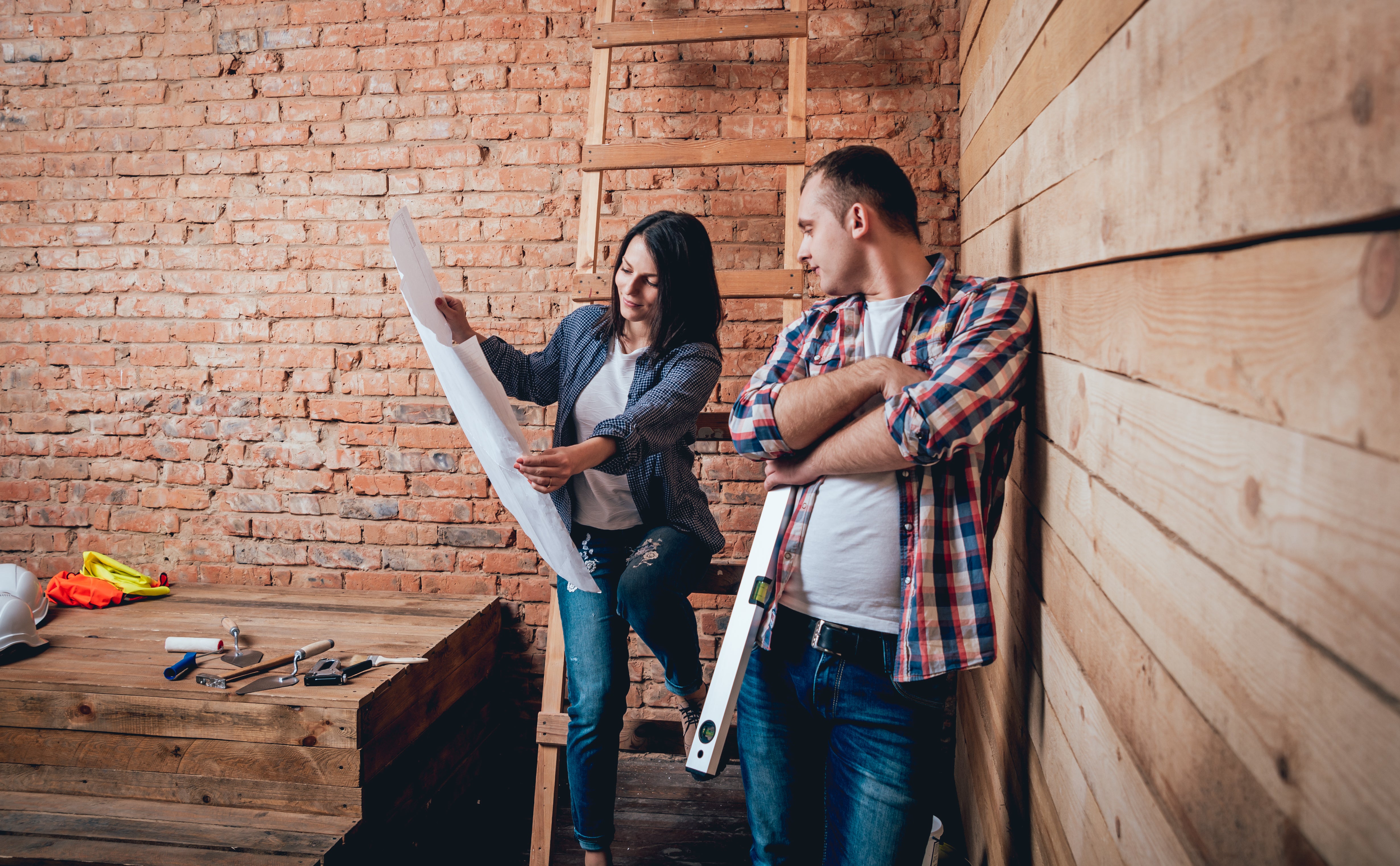 A woman and a man discuss a blueprint in a rustic, unfinished room with wooden walls and a brick wall. The woman holds and points at the blueprint, while the man, holding a level, listens. Tools are scattered on a wooden table nearby.