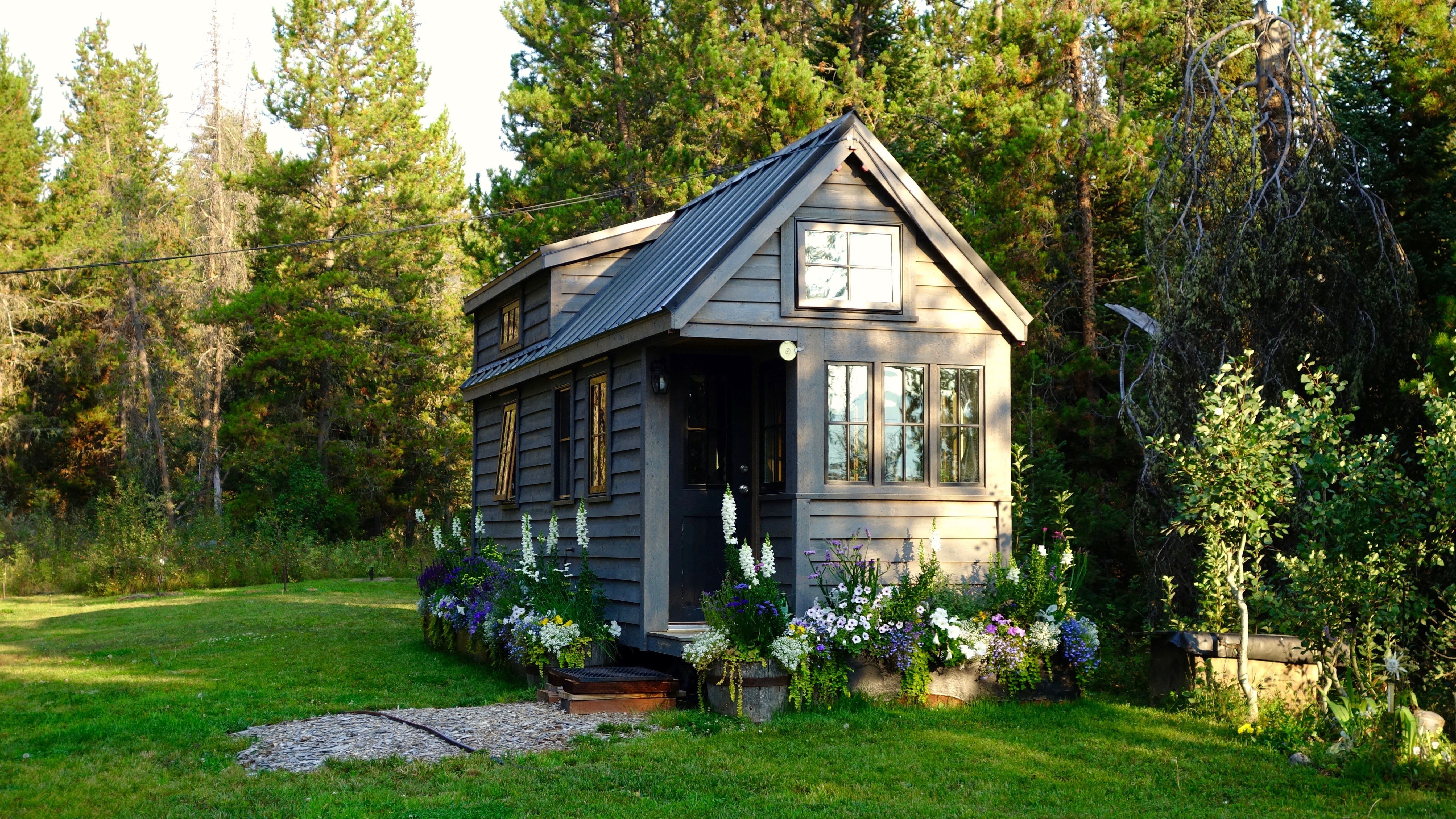 A small, gray tiny house with a sloped roof sits on a grassy lawn, surrounded by tall pine trees and colorful flower beds under bright sunlight.