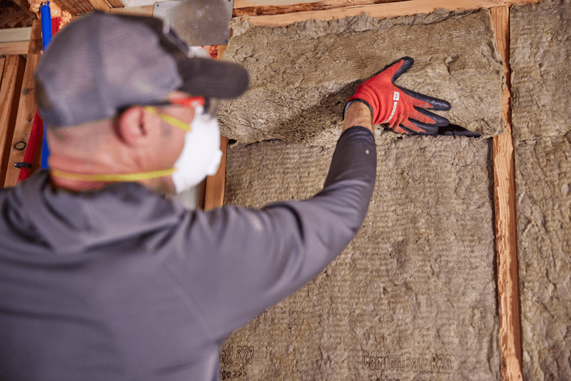 A man wearing a gray long-sleeve shirt, a white face mask, safety glasses, and red-and-black work gloves installs or adjusts insulation material between wooden wall framing boards.