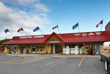 A lumber store with a red roof and several Canadian and blue flags on top. The sign reads TURKSTRA LUMBER. The storefront displays various building materials and the parking lot is mostly empty.